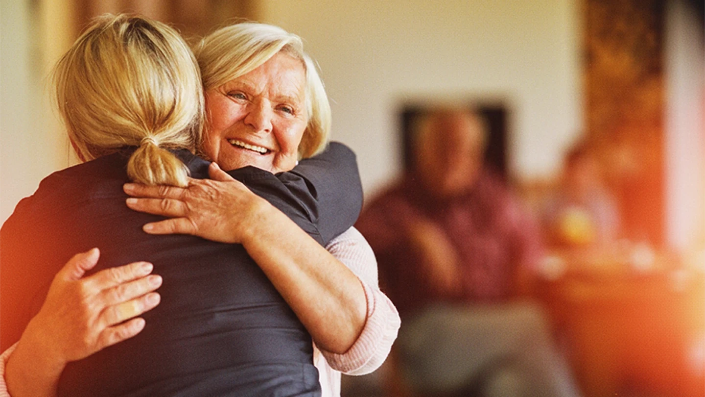 Eine &auml;ltere Frau und eine j&uuml;ngere Frau umarmen sich herzlich und l&auml;cheln. Im Hintergrund sind weitere Personen in einem warm beleuchteten Innenraum zu sehen.